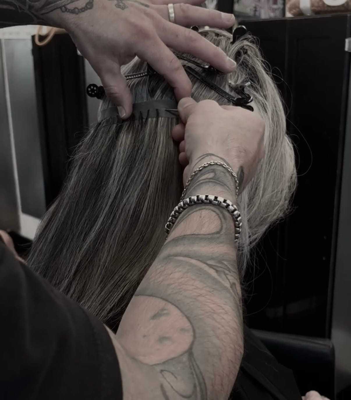 Tattooed stylist applying hair extensions to a person with blonde hair.